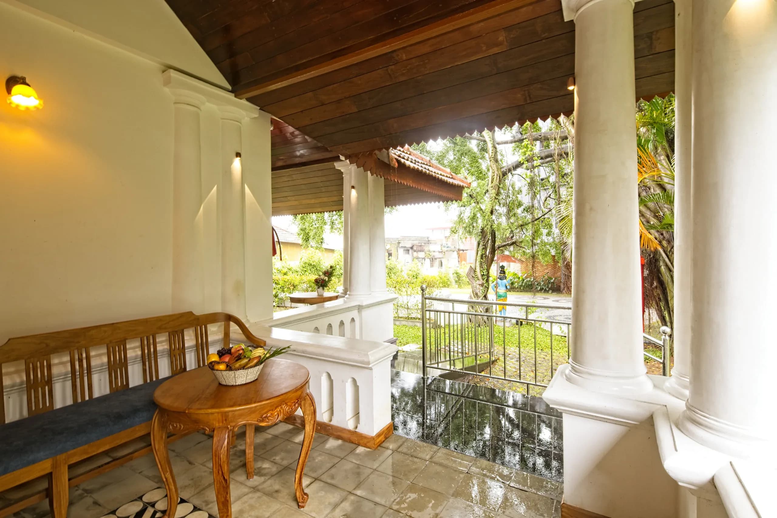 A pillared verandah with a carved teak bench and a small round table, open to the garden beyond.