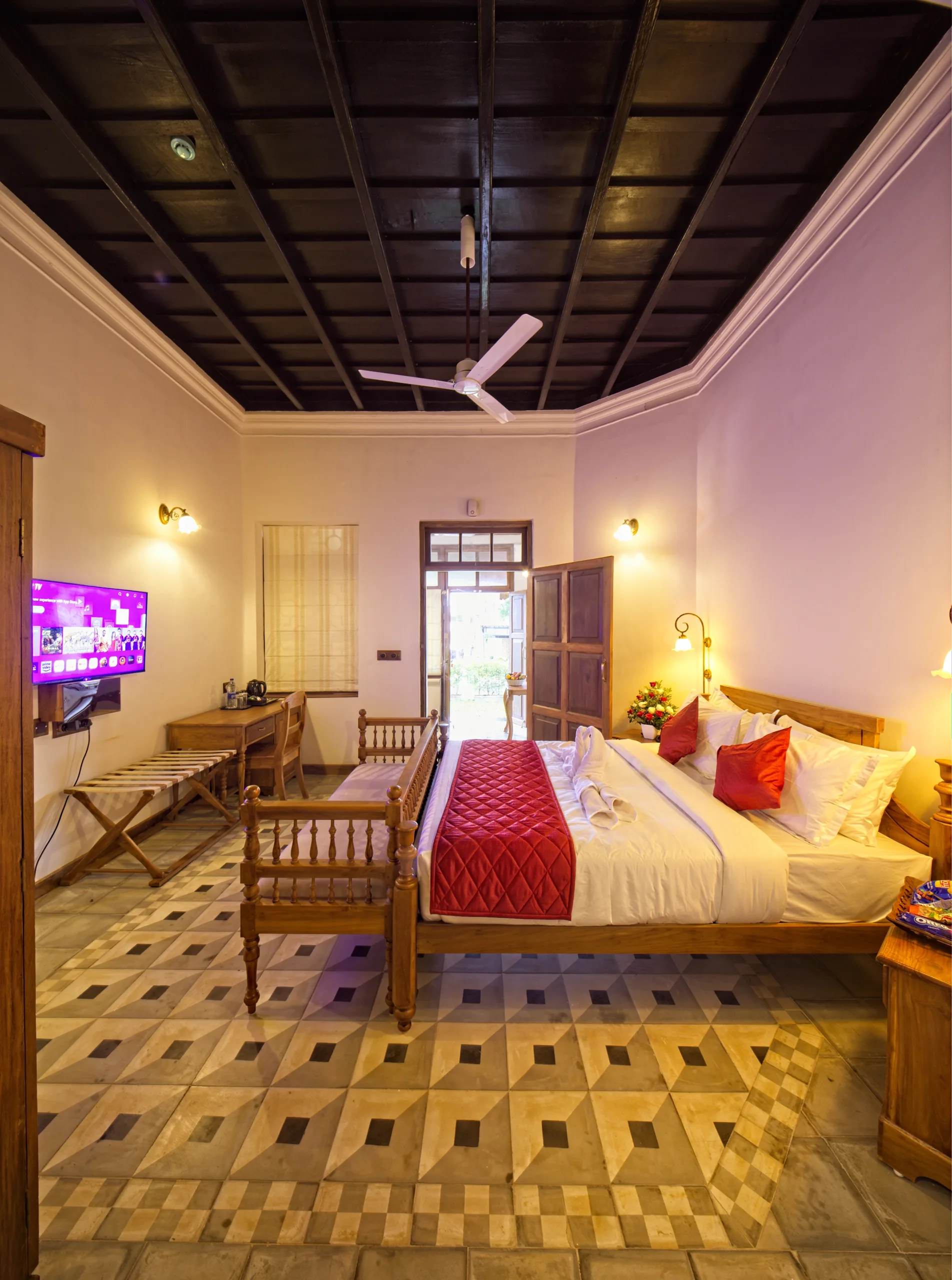 A high-ceilinged guest room with dark coffered timber above, a carved teak bed, a writing desk and a patterned tile floor in ochre and grey.