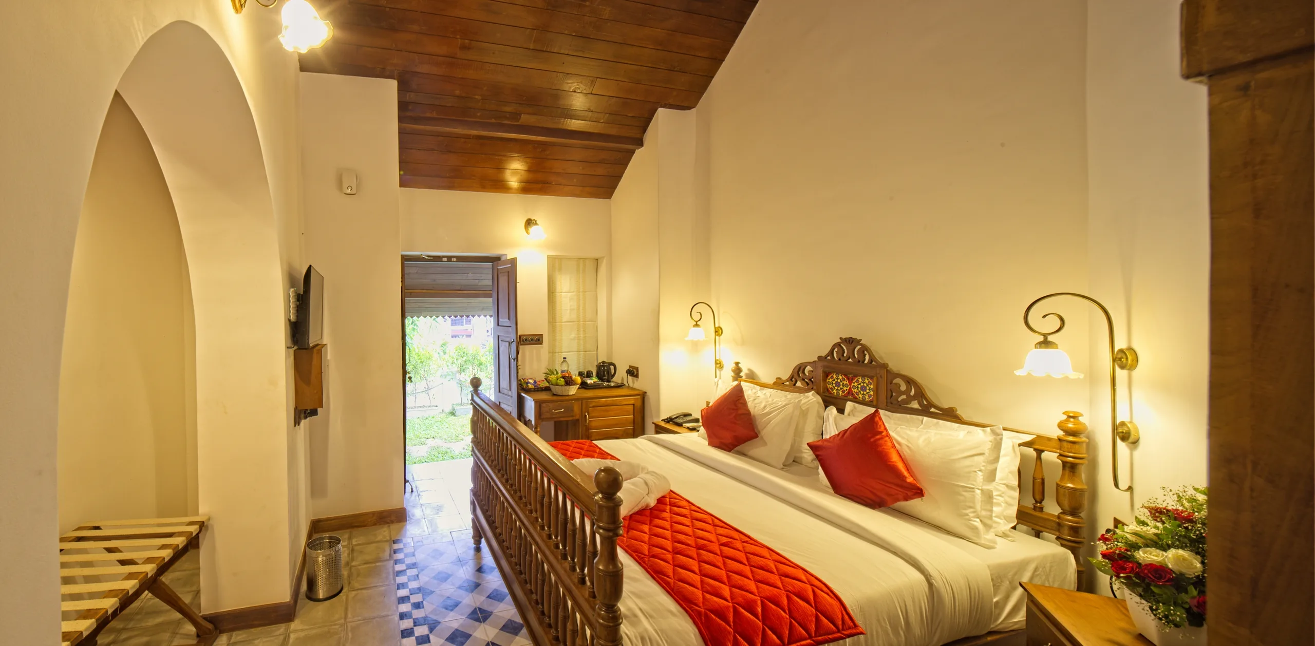 A guest room with a carved wooden bed under a polished timber ceiling, an arched doorway to one side and a door opening onto the garden.