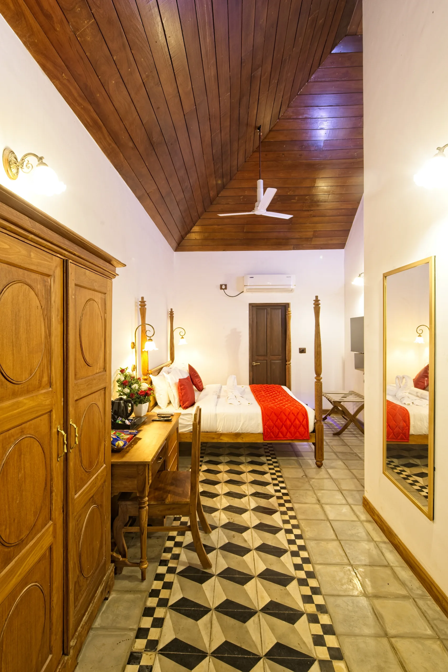 A guest room seen from the doorway, with a pitched wooden ceiling, a tall teak wardrobe, a writing desk and a black and white tile runner leading to the bed.