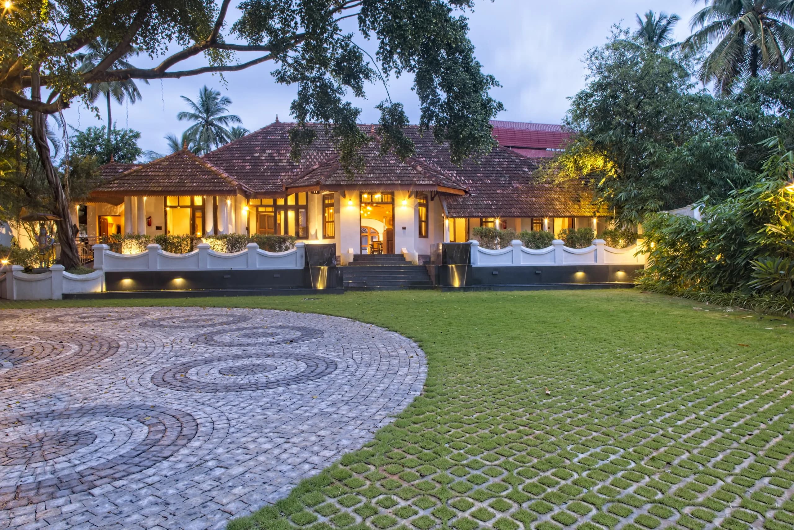 The bungalow lit from within at dusk, its windows glowing warm against a blue evening sky, seen across the lawn and cobblestone courtyard.