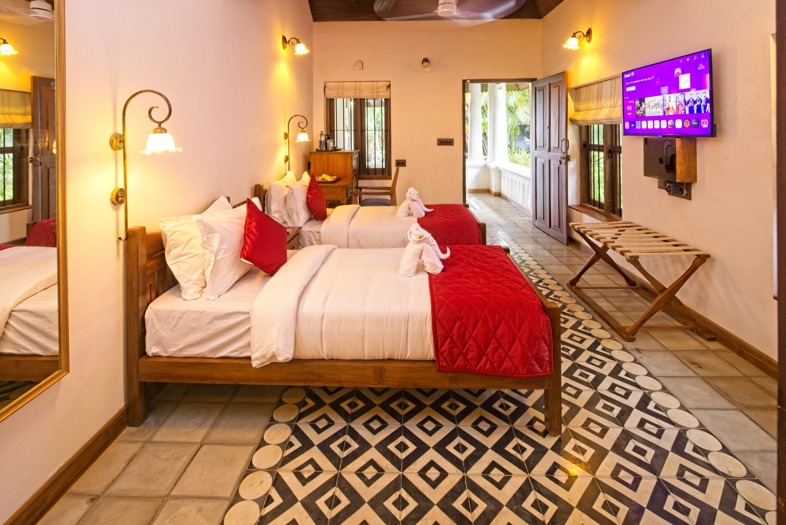 A twin guest room with two wooden beds, brass reading lamps and a patterned tile floor, with a door open to the greenery outside.