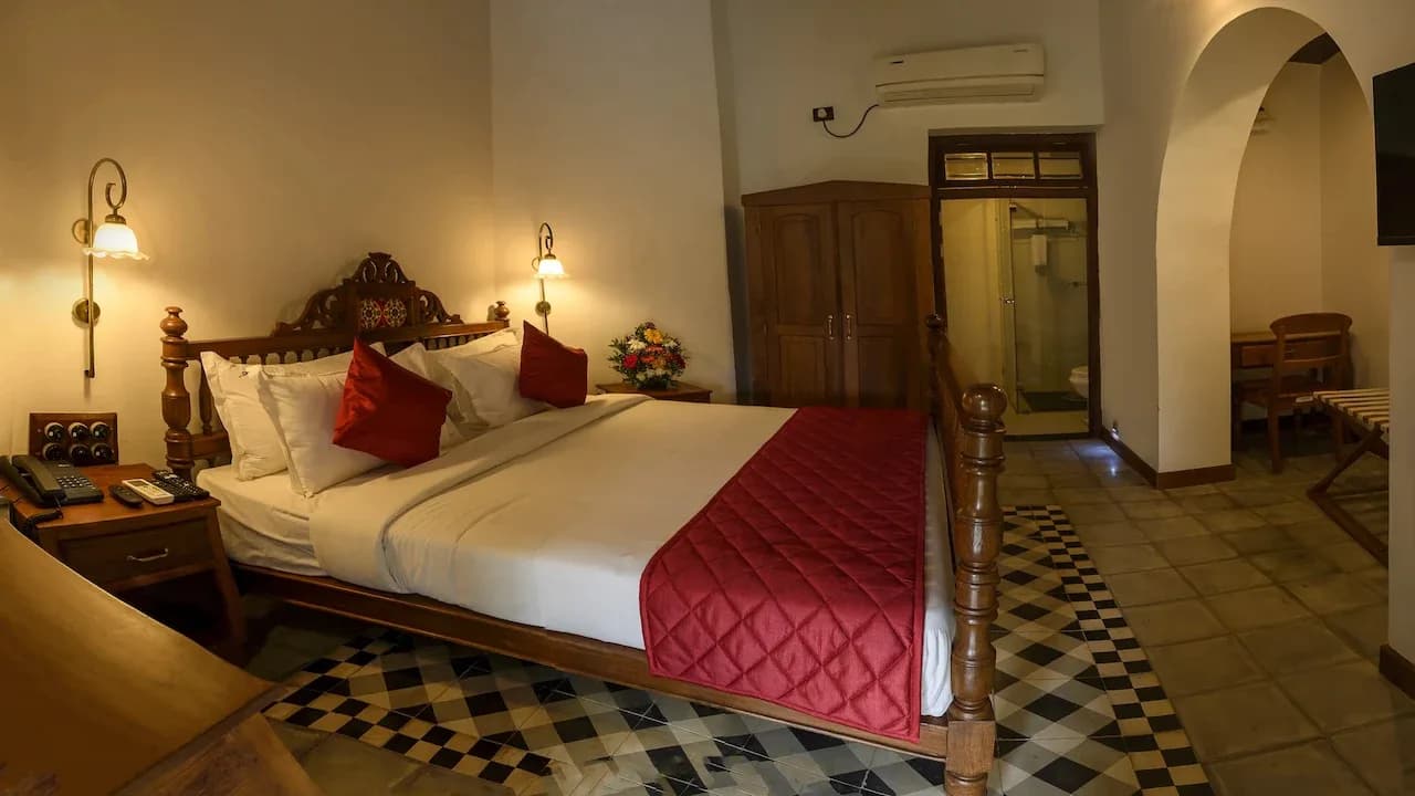 A guest room with a carved teak bed, brass wall lamps, a wooden wardrobe and a black and white chequered tile floor, with an arch through to a writing desk.