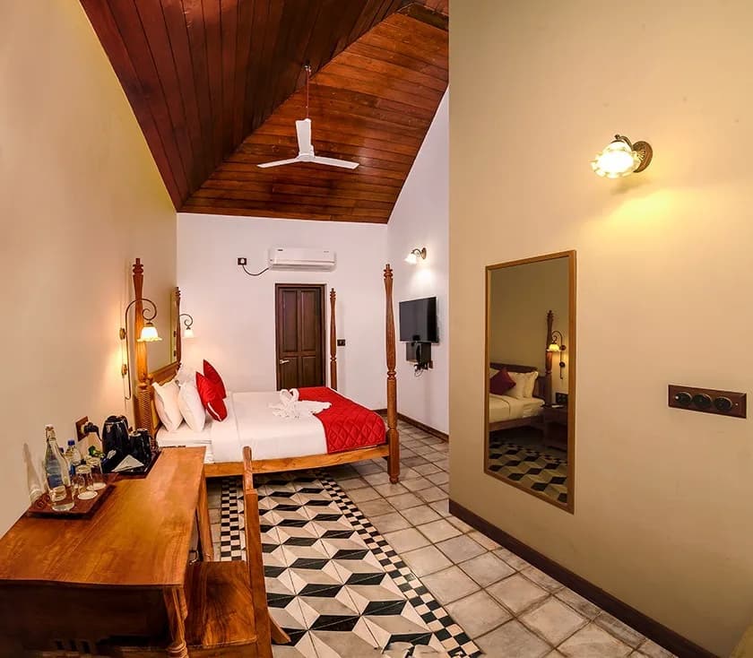 A guest room under a pitched timber ceiling, with a teak four-poster bed, a writing desk and a black and white tile runner leading to the door.