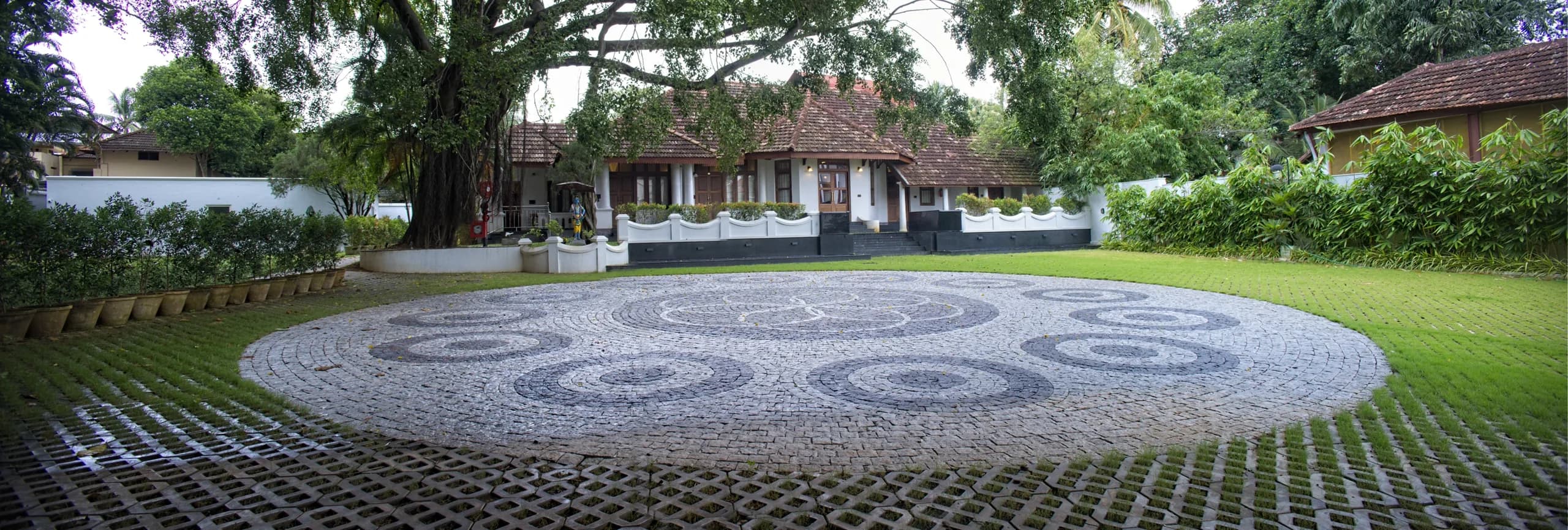 The bungalow's tiled roof and verandah behind the circular stone courtyard, framed by the branches of an old tree.