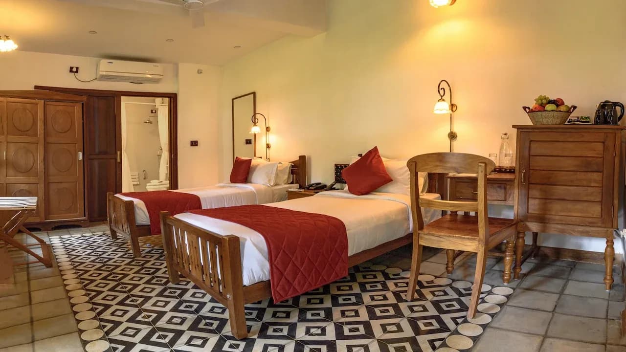 A twin guest room with two wooden beds under red quilted runners, brass wall lamps, a carved teak cabinet and a black and white geometric tile floor.