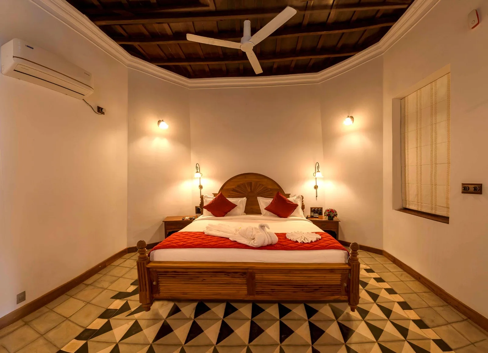 A guest room under a dark timber ceiling, with a carved sunburst headboard, brass wall lamps either side and a black and white triangle tile floor.