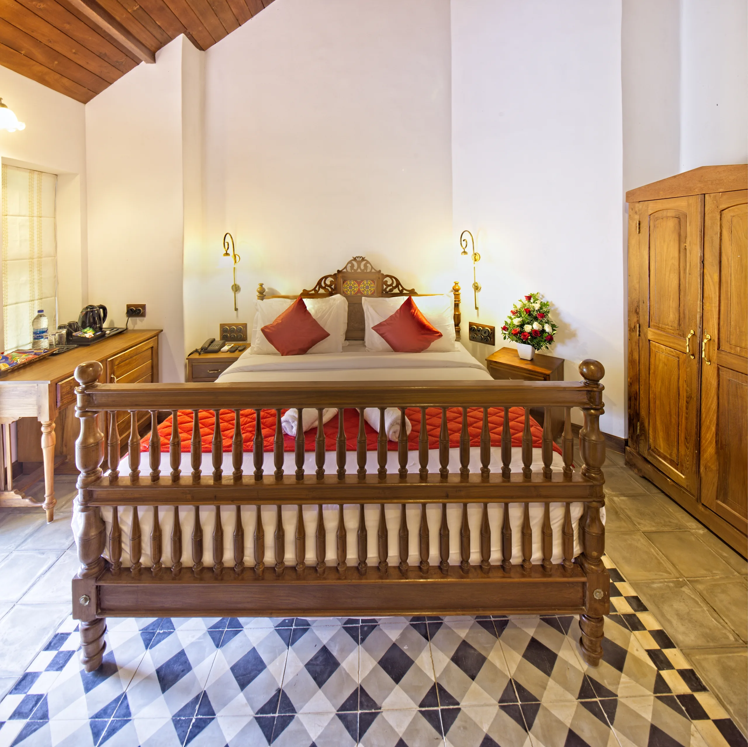 A guest room with a carved teak four-poster bed, brass wall lamps, a wooden wardrobe and a blue and white geometric tile floor.