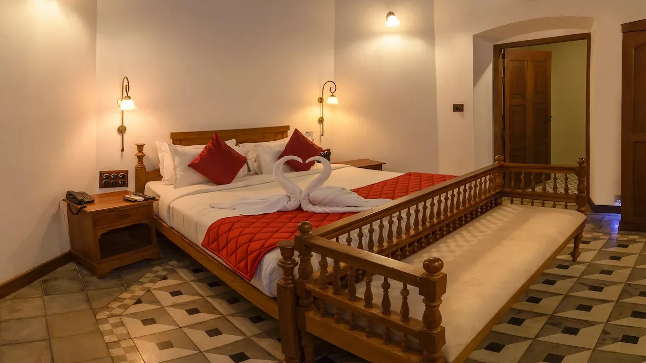 A guest room with a teak bed, brass wall lamps and a carved wooden day bed with a turned balustrade at its foot, over a patterned tile floor.
