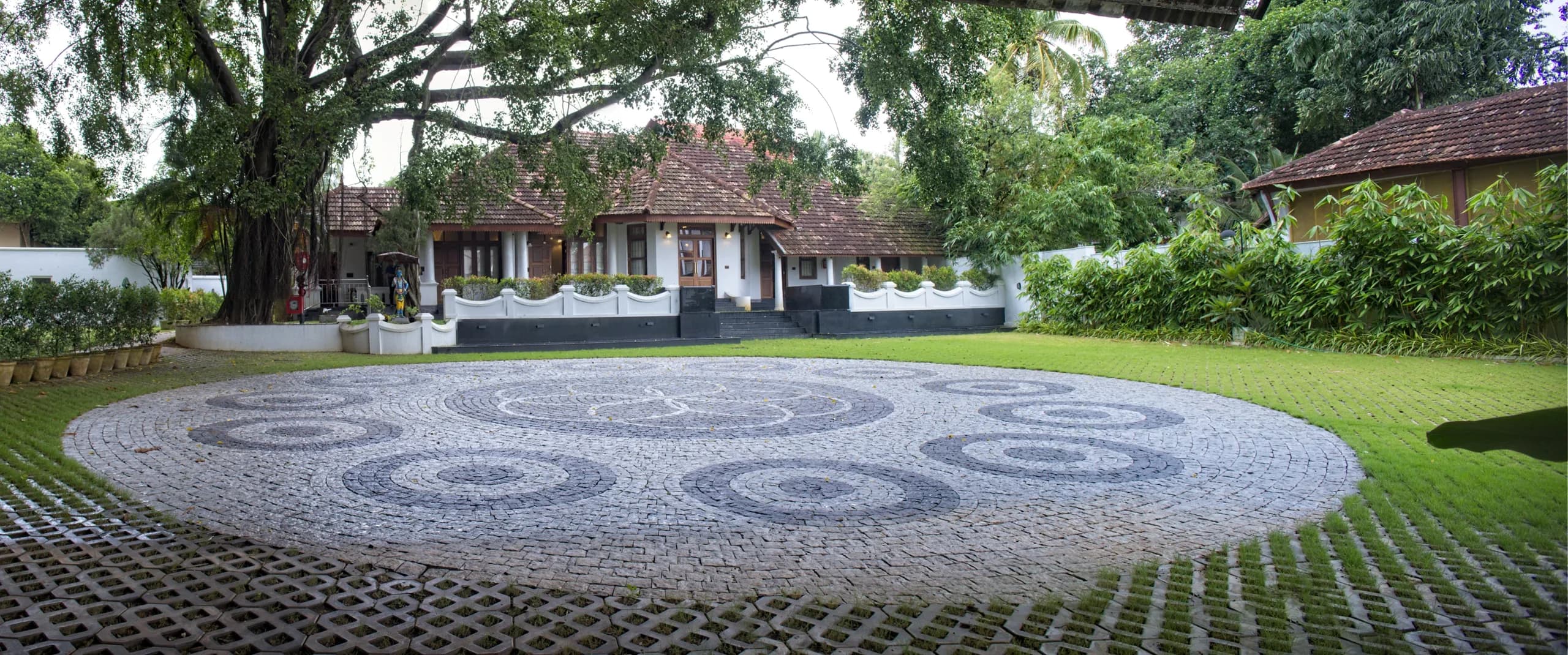 The heritage bungalow seen across the lawn, with a circular cobblestone courtyard laid in a spiral pattern under a large shade tree.